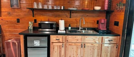 A compact kitchen with wooden cabinets, a stainless steel refrigerator, a microwave, a sink, and hanging pendant lights.