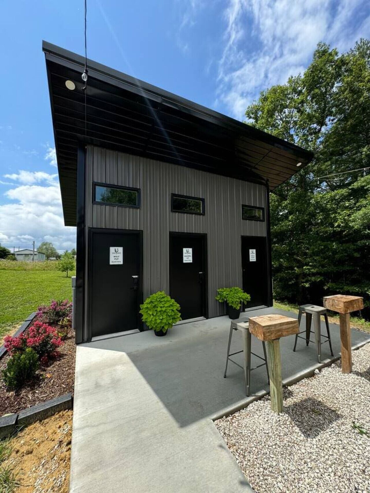A modern building with a black facade, three doors, and a small garden area with potted plants and a gravel path.