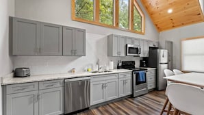 A modern kitchen with grey cabinets, a stainless steel dishwasher, a microwave, and a refrigerator. There is a wooden ceiling and large windows providing natural light.