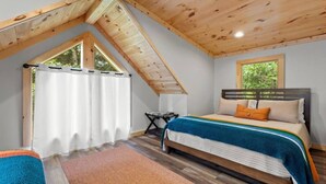 A bedroom with a sloped ceiling, a bed with a headboard, a window with curtains, and a small table.