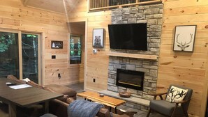 Ein gemütliches Blockhaus-Interieur mit Kamin aus Naturstein, Holzwänden, einem darüber montierten Fernseher und einem Essbereich mit Tisch und Stühlen.