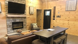 Ein gemütliches Blockhaus-Interieur mit Kamin aus Stein, einem flachbildfernseher darüber, einem Holzesstisch mit Barhocker und einer Ledersofa.