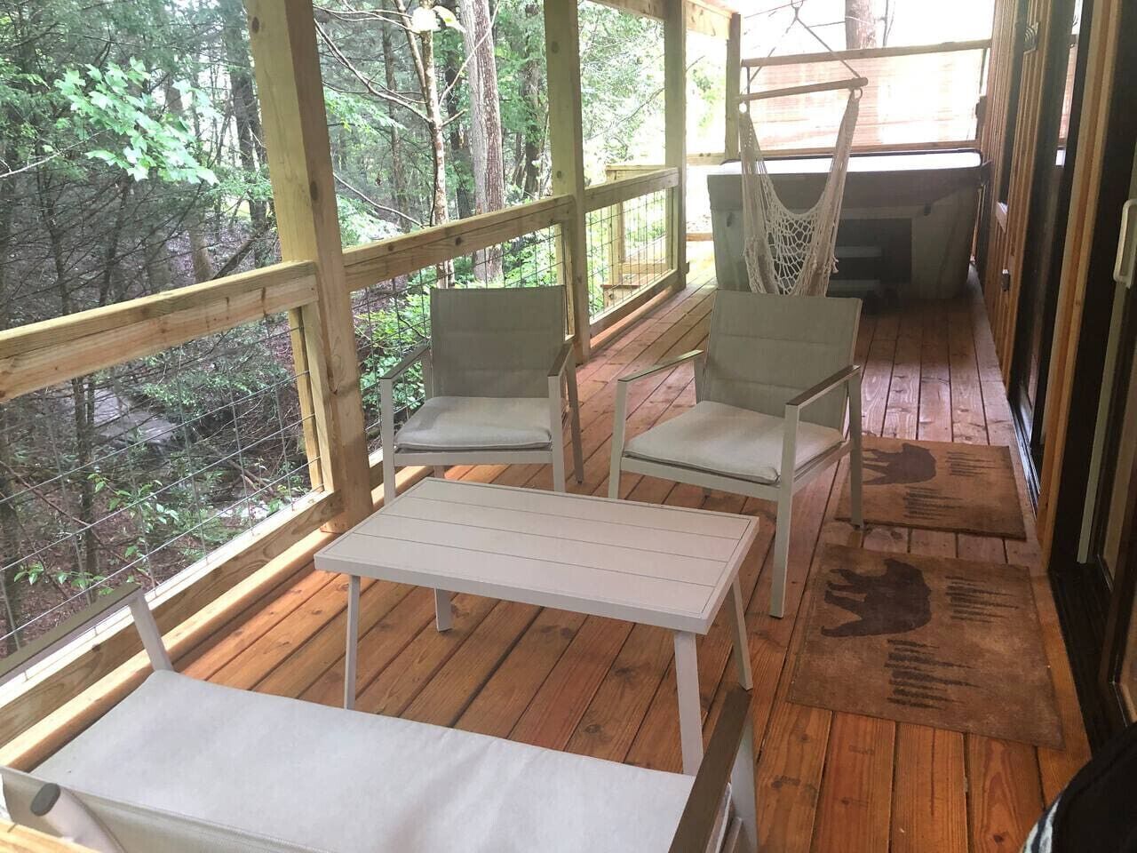 A wooden deck with two white tables and two chairs, surrounded by a wire mesh fence, overlooking a forested area.