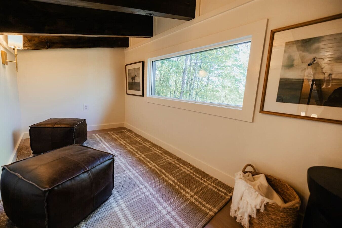 A room with a large window, a rug, two leather ottomans, a framed picture, and a woven basket.