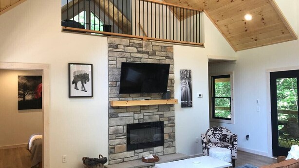 A spacious living room with a stone fireplace, a television mounted on the wall, and a high ceiling with exposed wooden beams.
