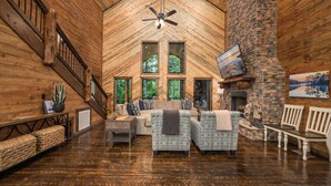 A spacious living room with a high ceiling, wooden walls, a stone fireplace, and a large window overlooking greenery.