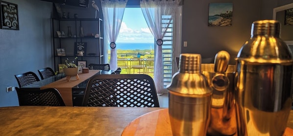 A dining area with a table and chairs, a view of the sea through a window, and a balcony with outdoor seating. - Ocean Views, Pool & Space for Groups (Vieques)