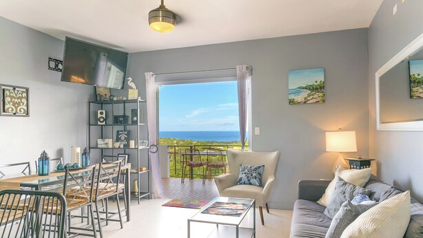 Un ampio soggiorno con balcone vista mare, una zona pranzo con tavolo e sedie e un angolo salotto accogliente con divano e cuscini.