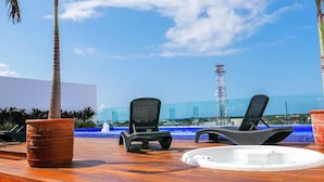 Een houten terras met twee loungesteilen en een jacuzzi, met uitzicht op een zwembad met een blauwe mozaïekrand en een hoge toren op de achtergrond.