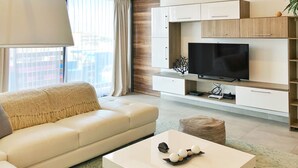 Een moderne woonkamer met een witte bank, een salontafel met decoratieve objecten, een flatscreen televisie op een houten kast en een groot raam met gordijnen.