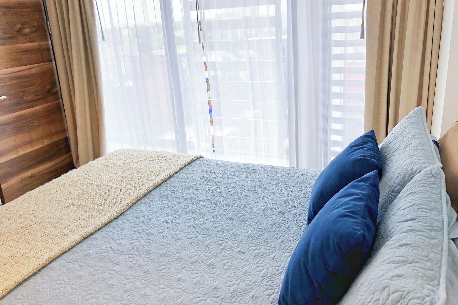 A bedroom with a bed, a window with blinds, and a view of buildings outside.