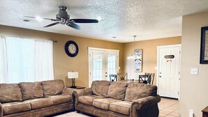 A living room with a brown leather sectional sofa, a dining table with chairs, a ceiling fan, a clock, and a door with a 'FAMILY' sign.