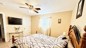 A bedroom with a bed, a ceiling fan, a TV, a desk, and a window with curtains.