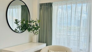 Una habitación moderna con un escritorio blanco, un espejo redondo, una planta verde, un sillón beige y un ventanal con cortinas translúcidas.