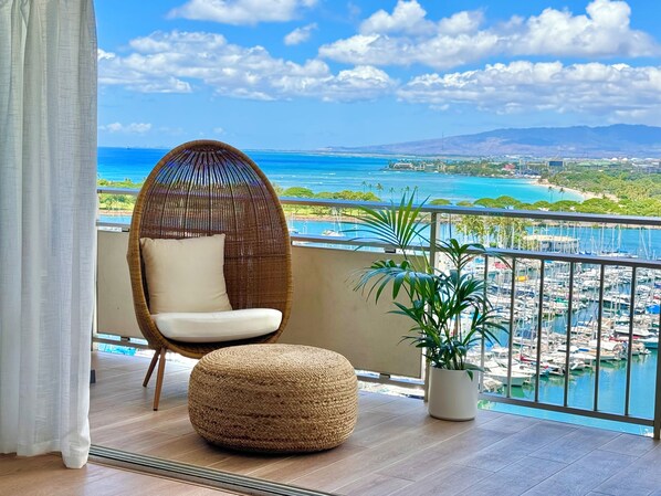 A balcony with a wicker chair, a round ottoman, and a potted plant.