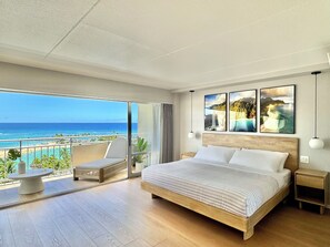 A spacious bedroom with a large bed, a balcony with a view of the beach, and artwork on the wall. - Oceanfront Dream Home (Car Available) (Honolulu)