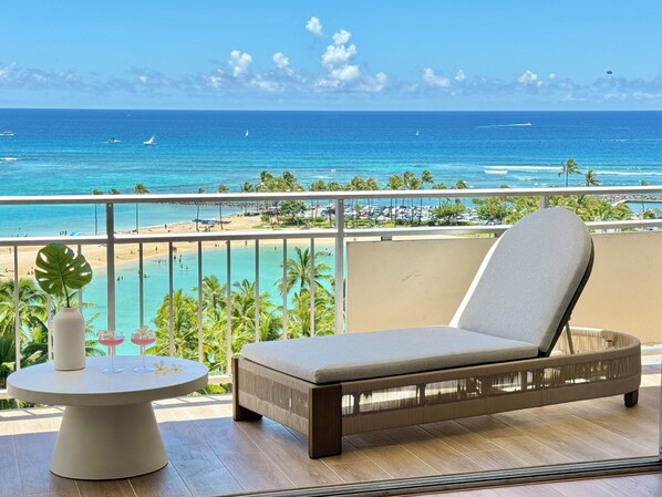 A balcony with a cushioned chair and a table, overlooking a beach and ocean with boats. - Oceanfront Dream Home (Car Available) (Honolulu)