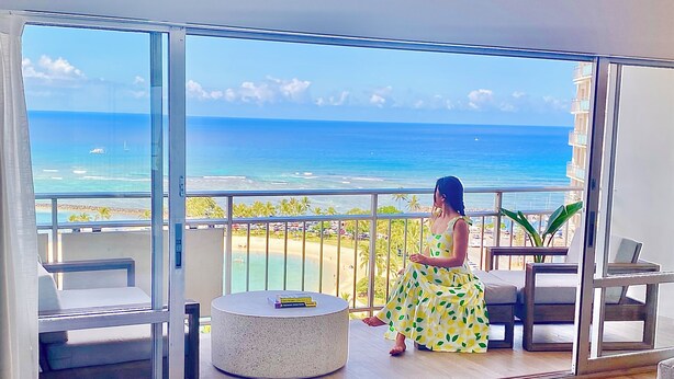 A balcony with a view of the ocean, a round table with a book, and a woman in a yellow dress.