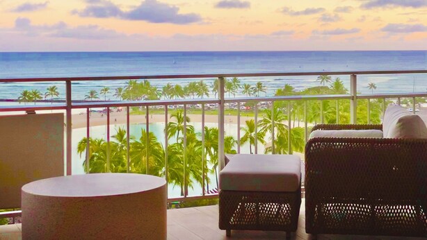 A balcony with wicker furniture, a round table, and a view of the beach and ocean. - Oceanfront Paradise (Car Available) (Honolulu)