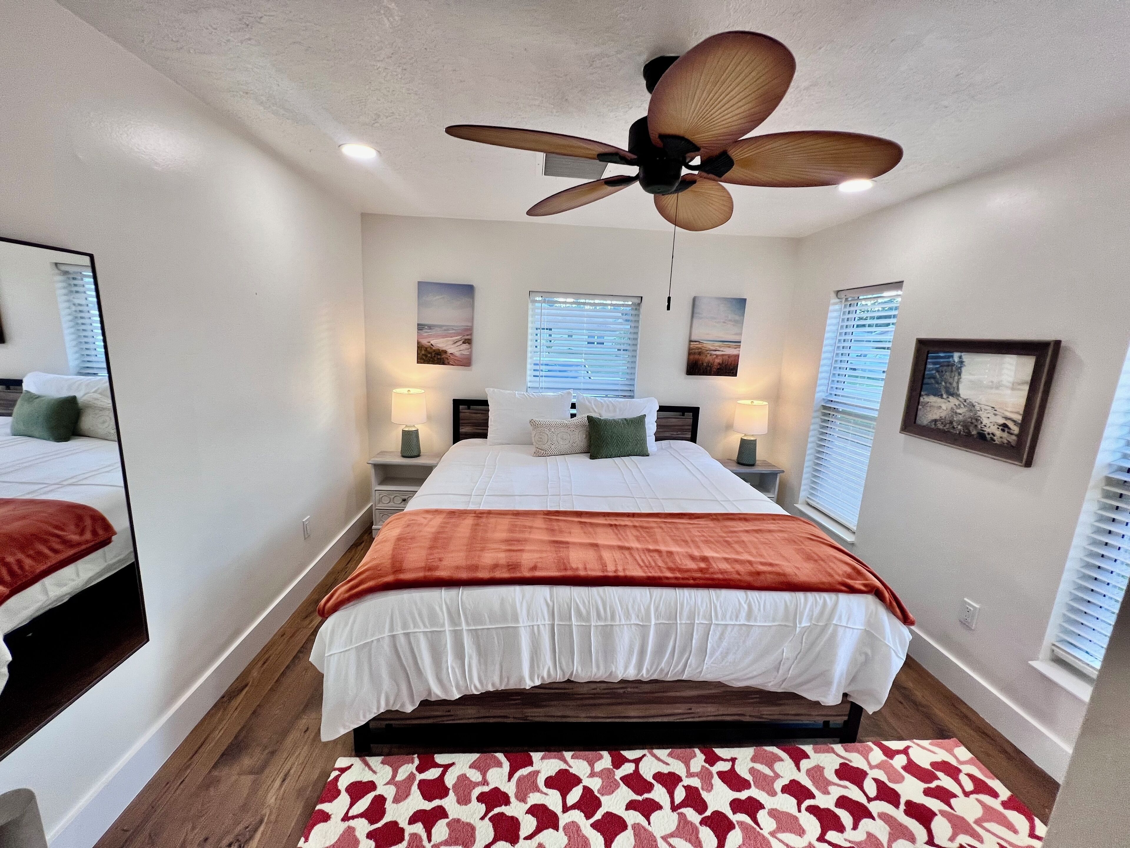 A bedroom with a bed, a ceiling fan, bedside lamps, a window with blinds, and a framed picture on the wall.