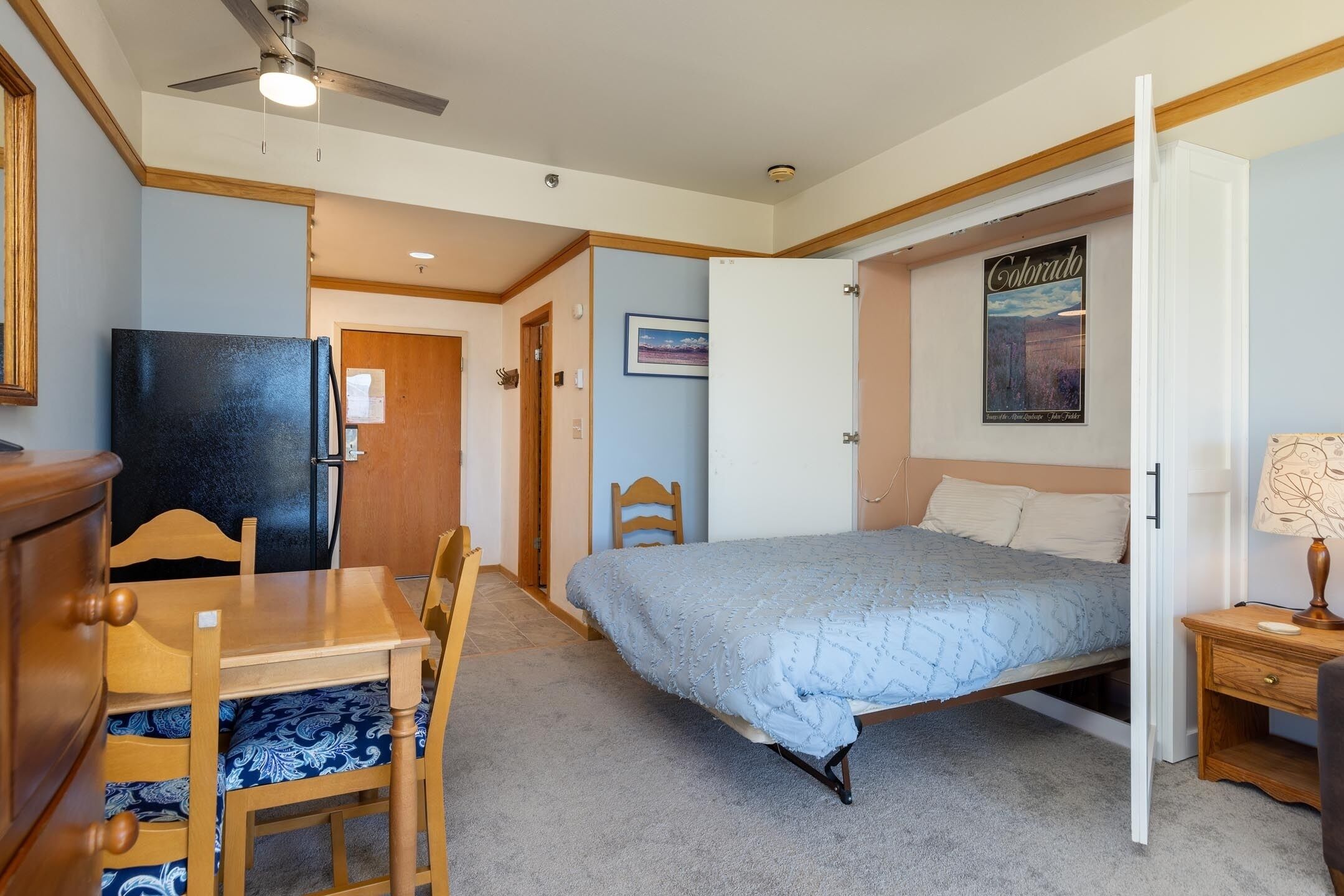 A bedroom with a bed, a desk, a chair, a refrigerator, a ceiling fan, and a door leading to another room.