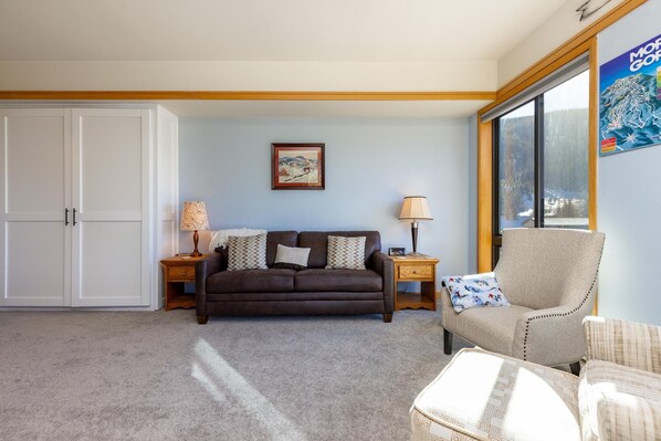 A living room with a brown sofa, a beige armchair, a wooden side table with a lamp, and a framed picture on the wall.