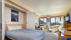 Habitación de hotel con una cama grande, un escritorio, una silla y vistas a las montañas a través de las ventanas.