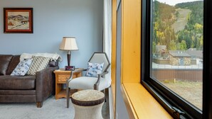 Un cuarto acogedor con un sofá de cuero marrón, un sillón blanco, una mesita de noche de madera y un taburete de mimbre. Una ventana ofrece vistas a un área boscosa y edificios.