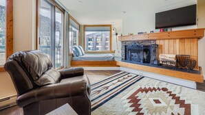 A modern living room with a leather sofa, a fireplace, a television, and large windows offering a view of buildings. - 1483 Lakeside 1 Bedroom Condo (Keystone)