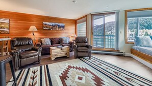 A cozy living room with a wooden wall, a sofa set, a coffee table, and a large window with a view of mountains. - 1483 Lakeside 1 Bedroom Condo (Keystone)