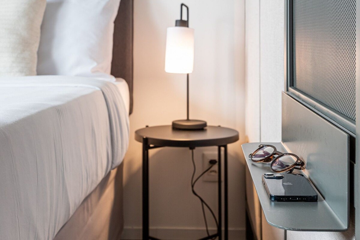 Ein Hotelzimmer mit einem Bett, einem Nachttisch mit Lampe, einer Brille und einem Smartphone.