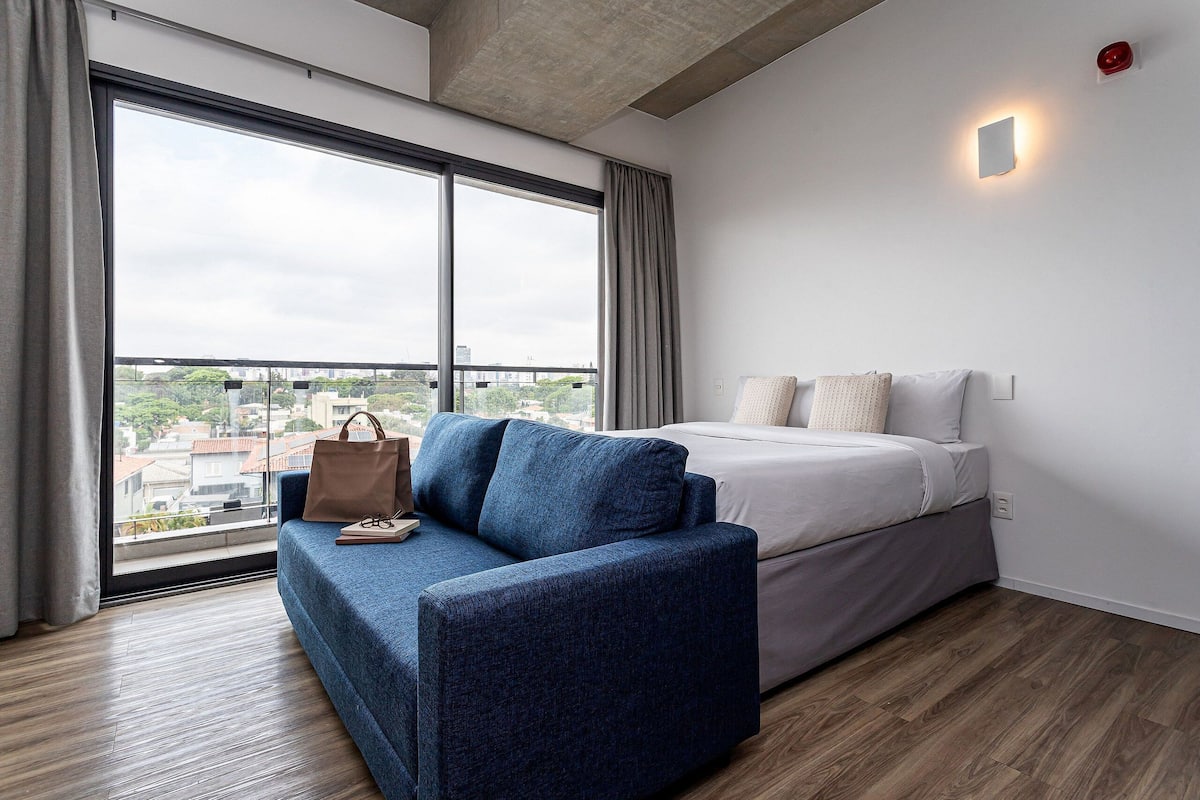 A modern hotel room with a large window, a sofa, a bed, and a view of the cityscape.
