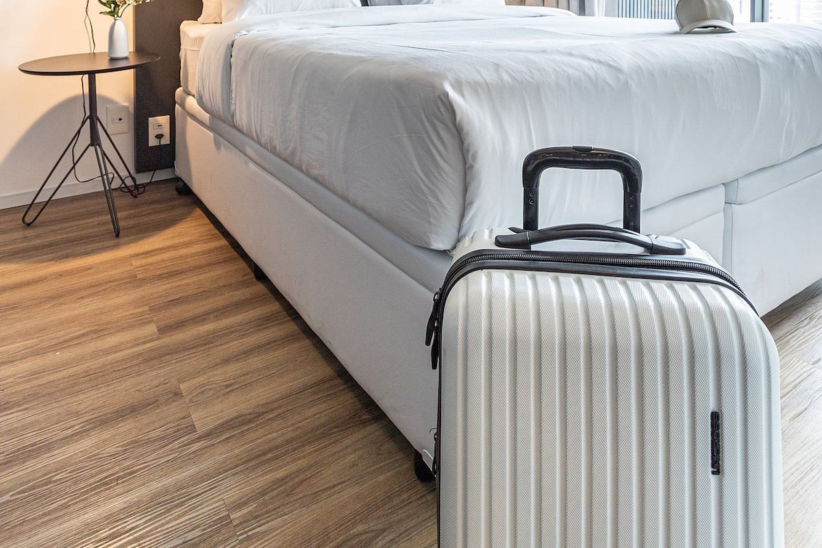 A modern hotel room with a large bed, a small bedside table with a lamp, a suitcase, and a view of the city through large windows.