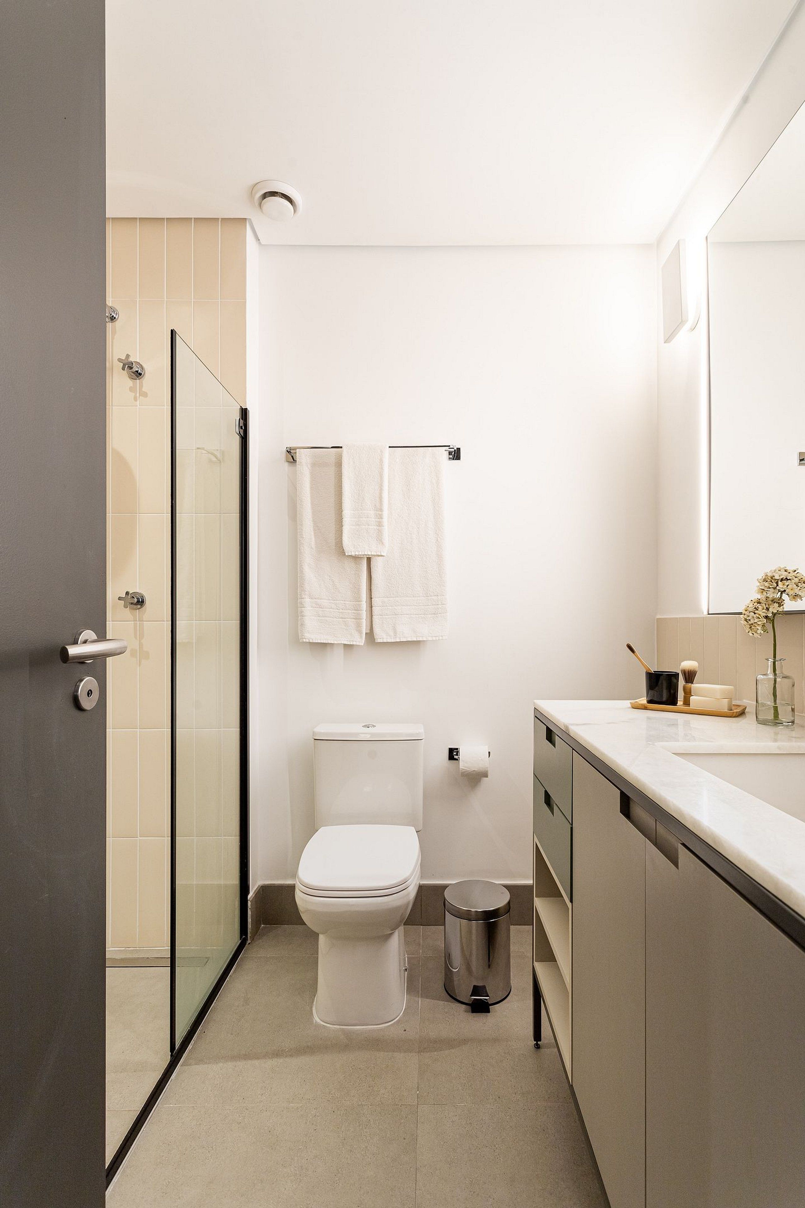 A modern bathroom with a toilet, a glass shower enclosure, a sink with a mirror, and a trash bin.
