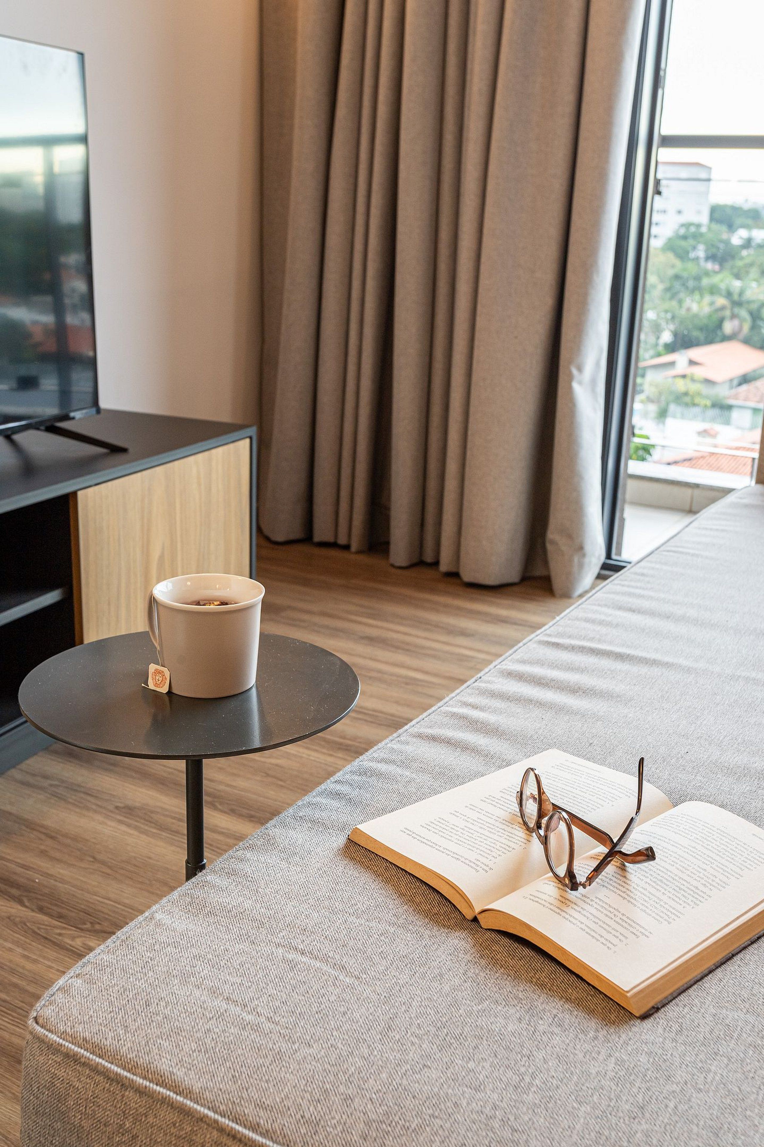 Une chambre d’hôtel moderne avec un canapé, une petite table ronde sur laquelle se trouvent une tasse et un sachet de thé, et un livre ouvert avec des lunettes posées dessus.