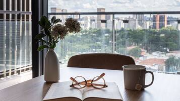 Una mesa con un libro, gafas, una taza, un jarrón con flores y vistas a un paisaje urbano a través de amplios ventanales.