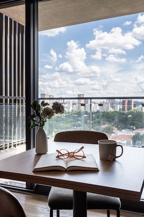 A table with a book, glasses, and a mug, a vase with flowers, and a view of a cityscape through large windows. - Apartamentos com vista incrível para São Paulo ao lado da Rebouças e Faria Lima - Pinheiros (São Paulo)