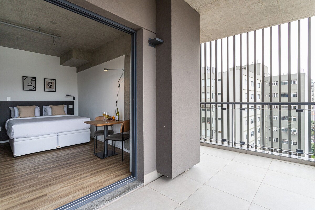 A modern hotel room with a bed, a small table, and a view of the city through a balcony.