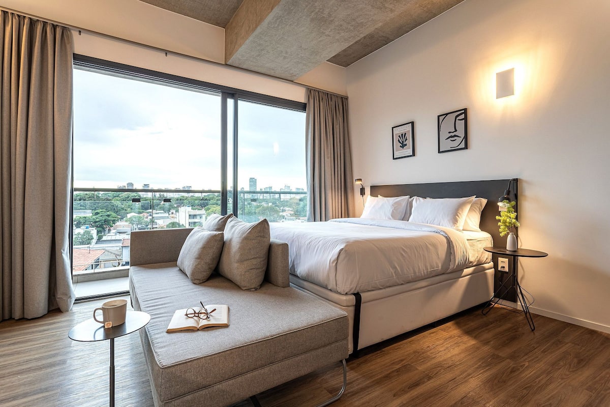A modern hotel room with a large bed, a sofa, a small table with a book and a mug, a nightstand with a lamp, and a city view.