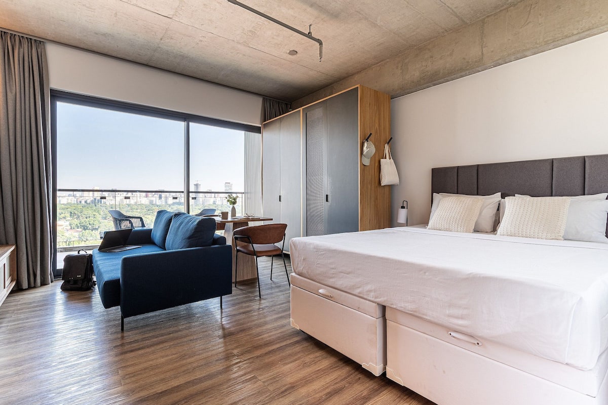 A modern hotel room with a large bed, a desk, a sofa, and a view of the city.