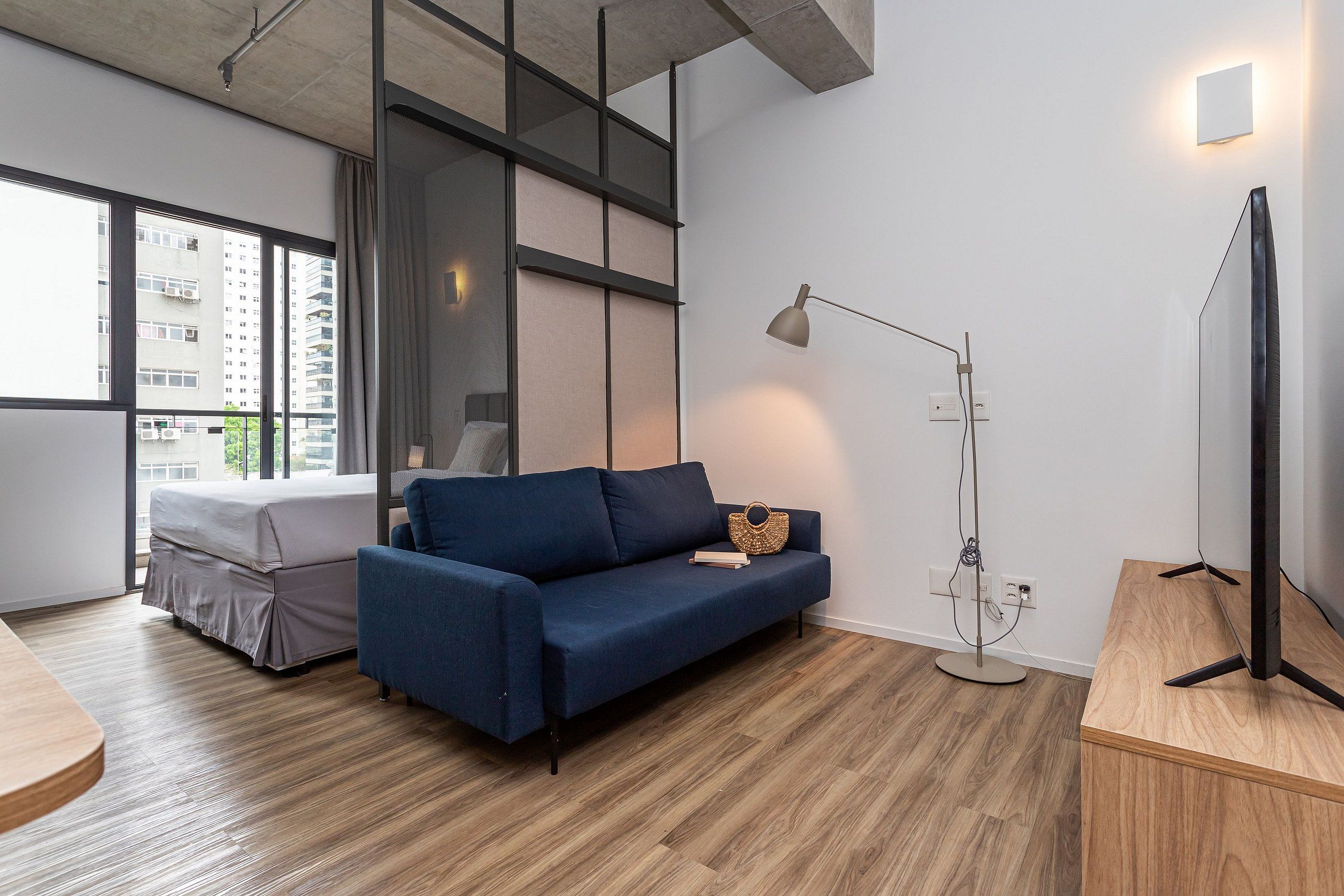 A modern room with a sofa, a bed, a standing lamp, and a view of buildings through a window.