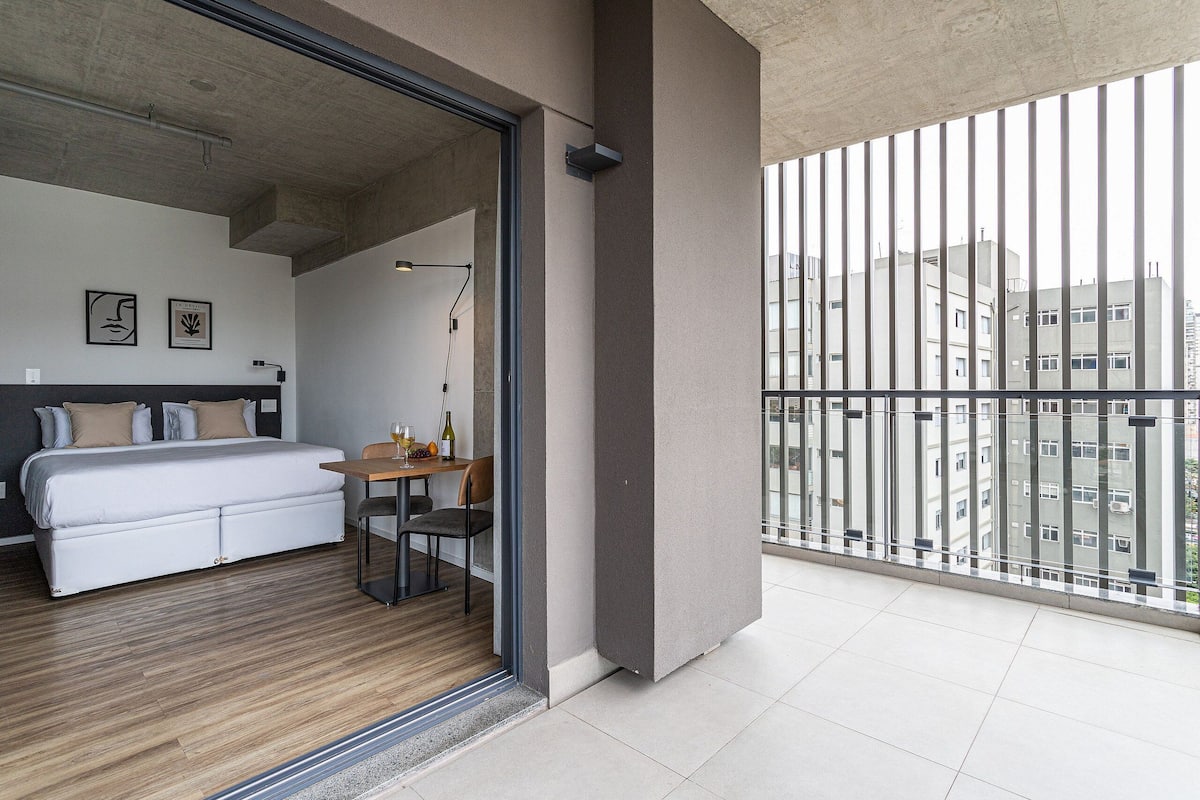 A modern hotel room with a bed, a small table, and a view of the city through a balcony.