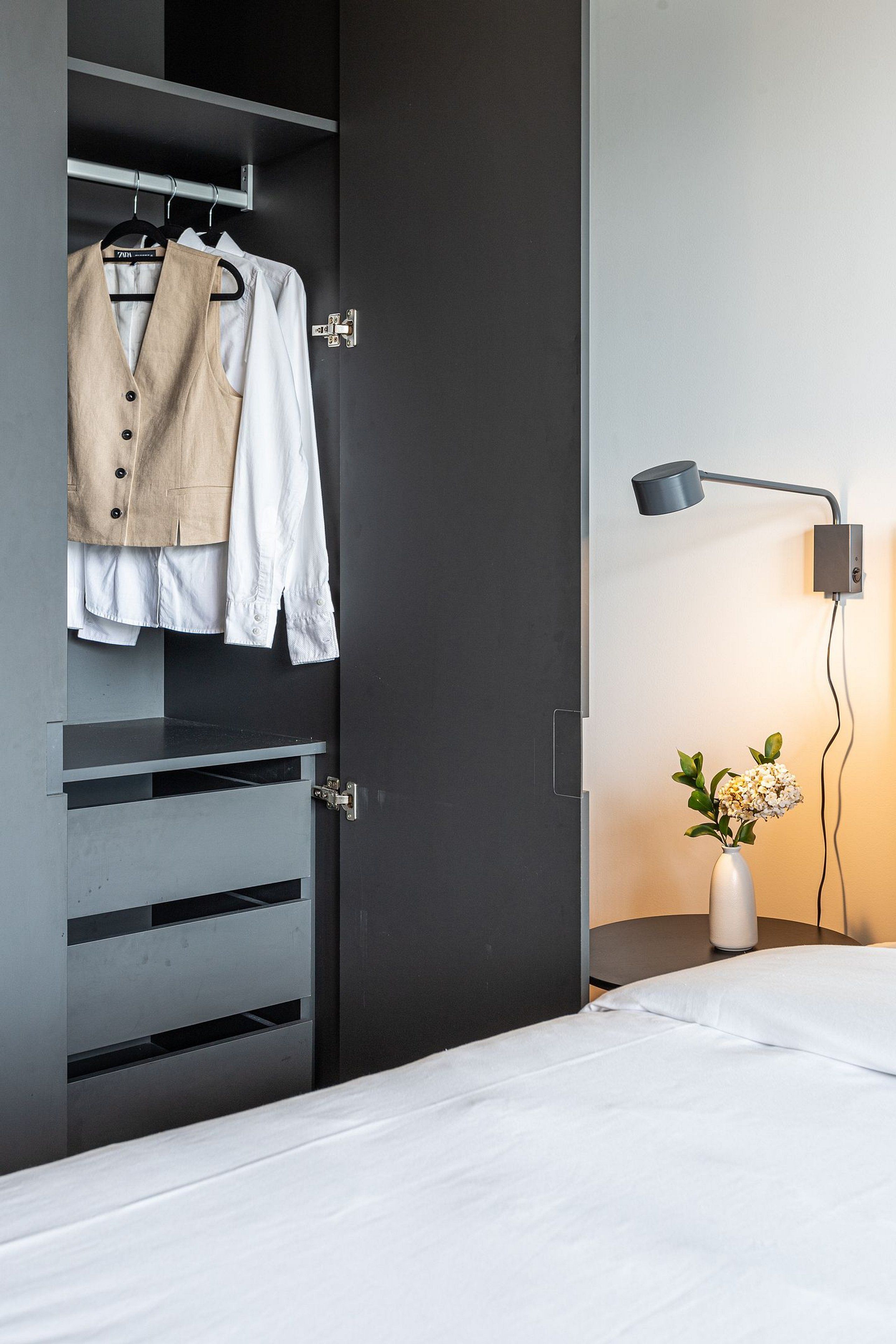Quarto com cama, guarda-roupa cheio de roupas, escrivaninha e luminária de mesa.