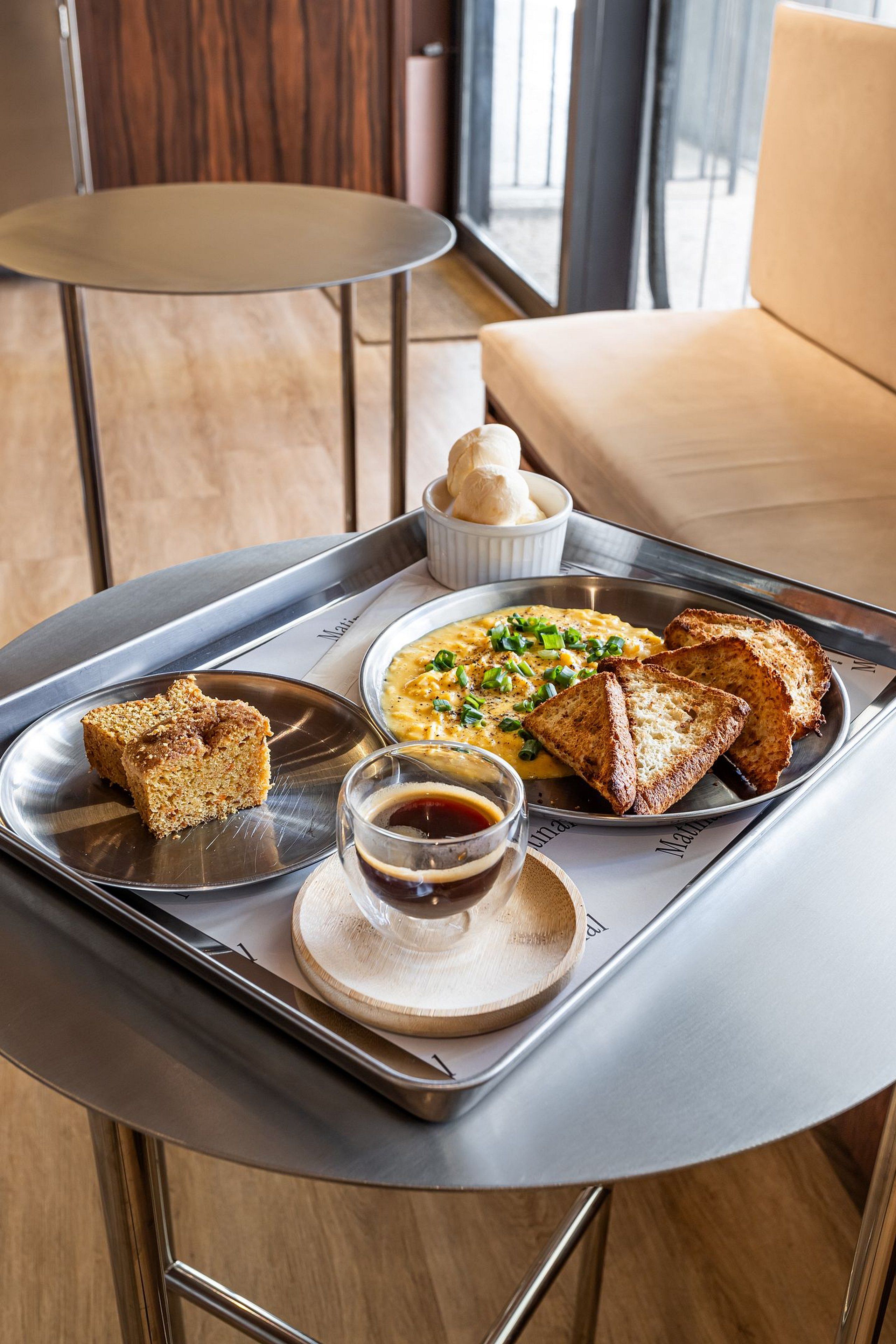 A breakfast tray with toast, scrambled eggs, and a small bowl of soup on a table in a modern room with a sofa and a window.