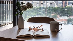 A table with a book, glasses, and a mug, a vase with flowers, and a view of a cityscape through large windows. - Apartamentos com vista incrível para São Paulo ao lado da Rebouças e Faria Lima - Pinheiros (São Paulo)