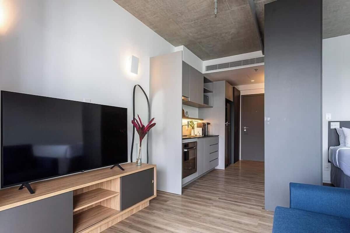 A modern hotel room with a flat-screen TV, a blue sofa, a wooden entertainment unit, and a kitchen area in the background.