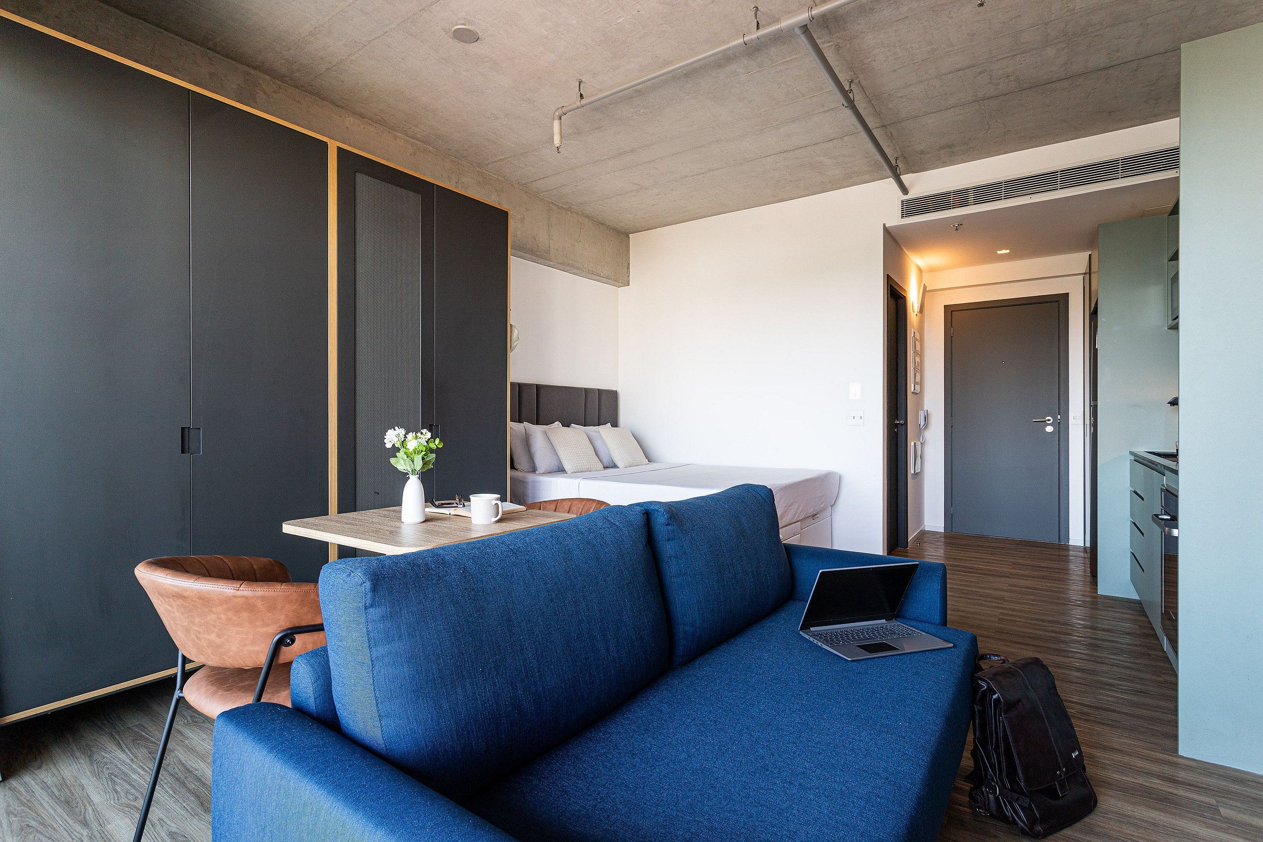 A modern hotel room with a blue sofa, a wooden dining table, a bed, and a kitchenette.