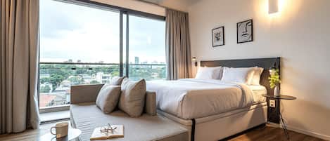 Ein modernes Hotelzimmer mit einem großen Bett, einer Couch, einem kleinen Tisch mit einem Buch und einer Tasse, einem Nachttisch mit einer Lampe und Blick auf die Stadt.