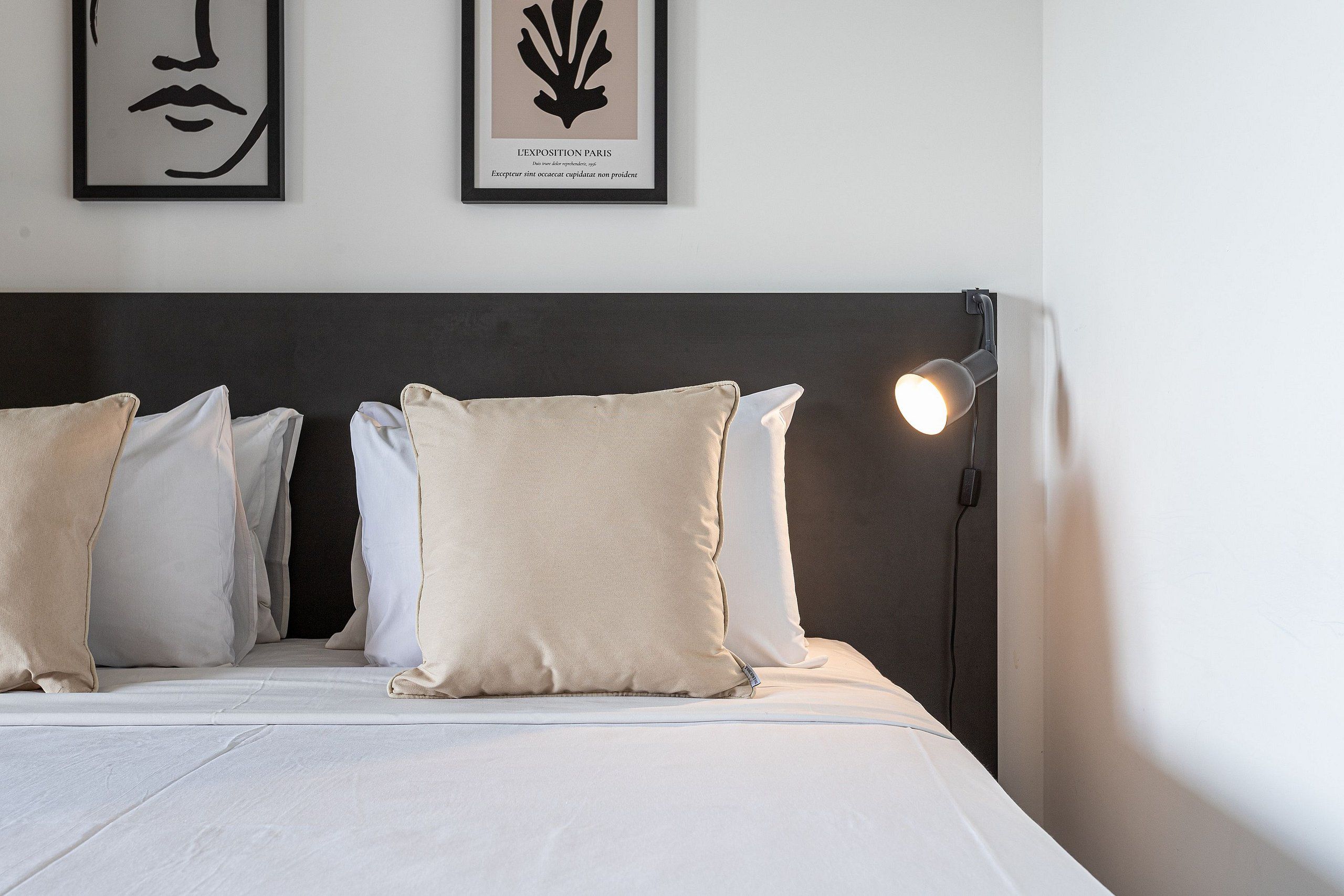 Un lit bien fait, avec du linge de lit blanc et des oreillers beige, une tête de lit sombre et deux œuvres d’art encadrées accrochées au mur.
