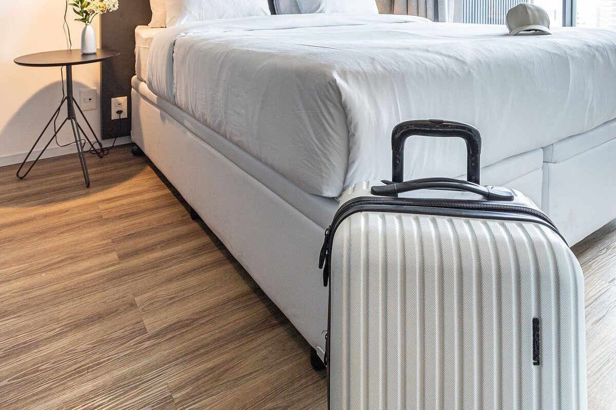 A modern hotel room with a large bed, a small bedside table with a lamp, a suitcase, and a view of the city through large windows.
